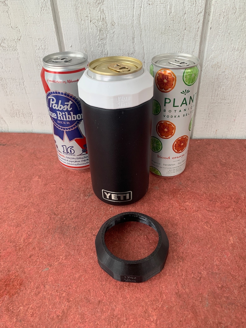 Op de afbeelding: Een zwarte YETI-blikjeskoeler met een witte bovenkant, naast drie drankblikjes. E&eacute;n blikje is een Pabst Blue Ribbon-bier en de andere is een Plant Botanical Vodka Seltzer. Een zwarte plastic ring ligt ervoor.