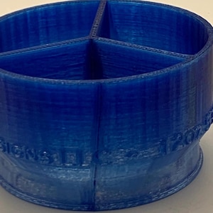 May include: A blue, 3D-printed organizer with four compartments. The cylindrical container has a wide opening and a base with embossed text. The design is a solid, vibrant blue color.