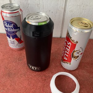 Op de afbeelding: Een zwarte Yeti-blikjeskoeler met een witte rand en een blikje erin. Er zijn ook twee bierblikjes, een Pabst Blue Ribbon en een Stella Artois. De koeler is gemarkeerd met "17oz Slim".