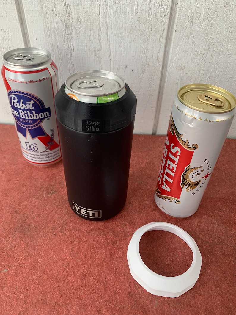 Op de afbeelding: Een zwarte Yeti-blikjeskoeler met een witte rand en een blikje erin. Er zijn ook twee bierblikjes, een Pabst Blue Ribbon en een Stella Artois. De koeler is gemarkeerd met "17oz Slim".