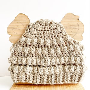 May include: A beige crochet hat with a textured, bobble stitch design. The hat is displayed on a wooden stand with heart-shaped cutouts. The hat is made of a soft, natural-colored yarn.