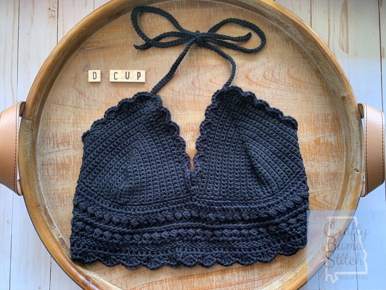 May include: Black crochet halter top with a scalloped edge and a D cup size label. The top is displayed on a wooden tray.