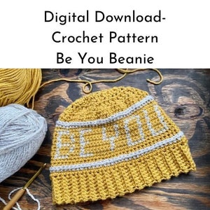 May include: A crocheted beanie in yellow with a white band and the words "Be You" in white lettering. The beanie is on a wooden surface.