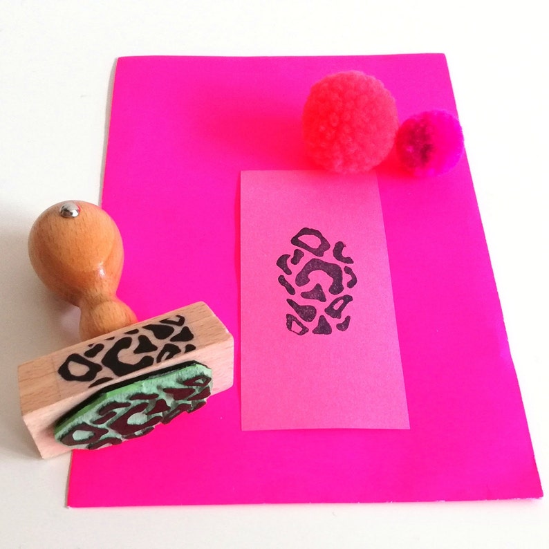 May include: A wooden stamp with a black and white leopard print design on a pink surface. The stamp is next to a pink sheet of paper with a black and white leopard print design stamped on it. There are two pink pom-poms on the surface.