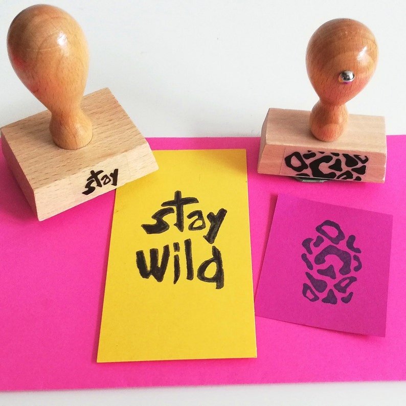 May include: Two wooden stamps with black ink. One stamp reads "stay wild" and the other has a leopard print design. The stamps are on a pink surface with two yellow and purple note cards.