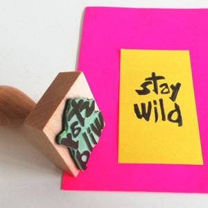 May include: A wooden stamp with the words "Stay Wild" in green and black ink. The stamp is on a pink surface next to a yellow card with the words "Stay Wild" written in black ink.