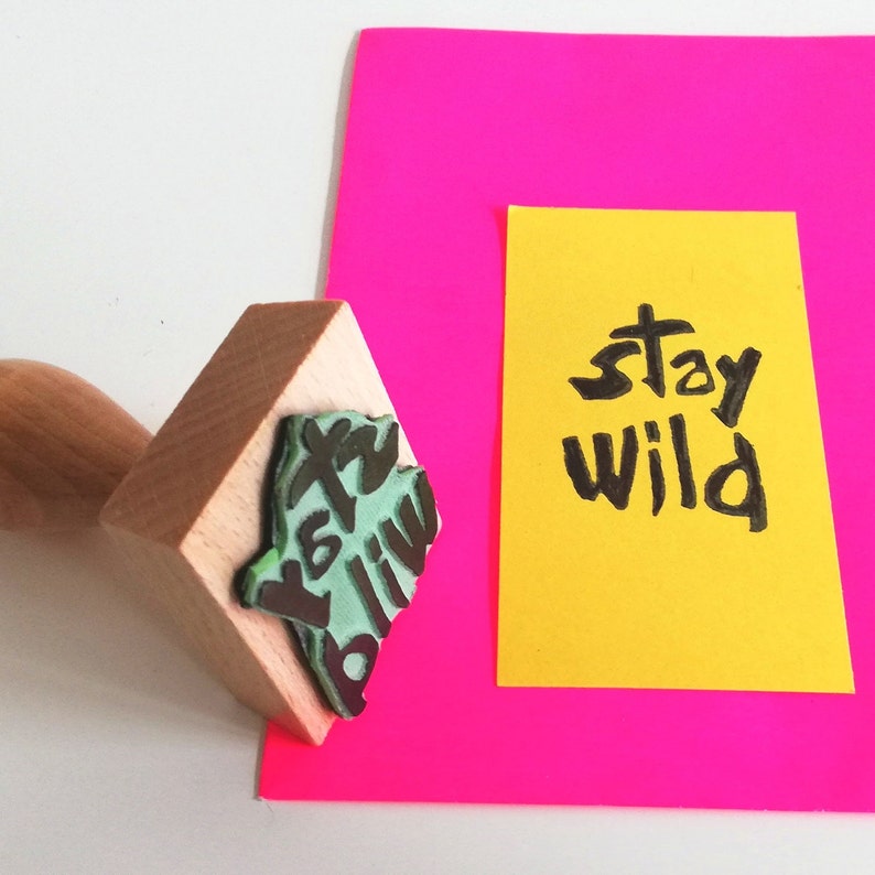 May include: A wooden stamp with the words "Stay Wild" in green and black ink. The stamp is on a pink surface next to a yellow card with the words "Stay Wild" written in black ink.