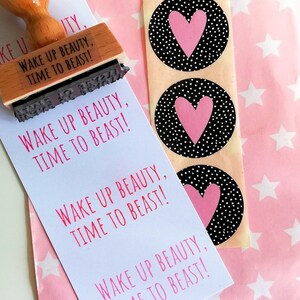 Puede incluir: Una tarjeta blanca con el texto "Wake Up Beauty, Time To Beast!" repetido cuatro veces. La tarjeta est&aacute; junto a una hoja de papel de regalo rosa con estrellas blancas y tres pegatinas blancas y negras con corazones rosas.