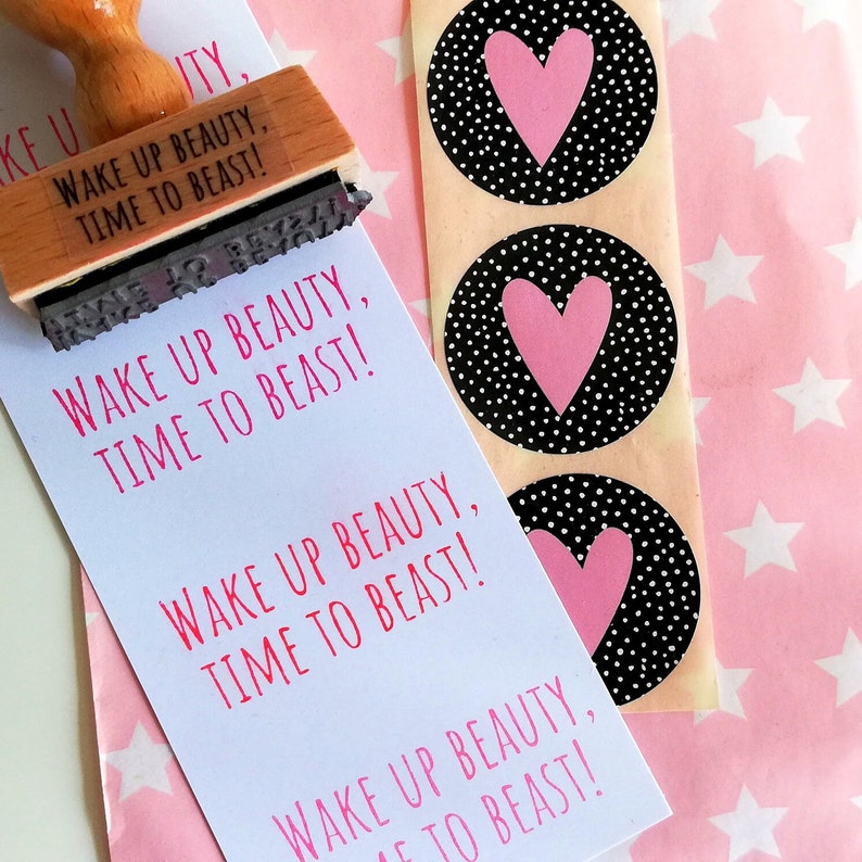 Puede incluir: Una tarjeta blanca con el texto "Wake Up Beauty, Time To Beast!" repetido cuatro veces. La tarjeta est&aacute; junto a una hoja de papel de regalo rosa con estrellas blancas y tres pegatinas blancas y negras con corazones rosas.