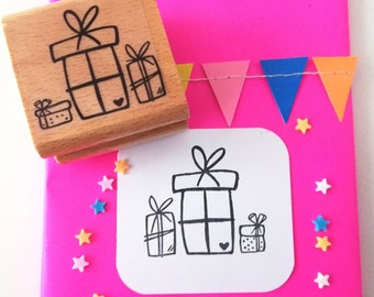 以下が含まれることがあります： 3つのギフトボックスのデザインが施された木製スタンプのクローズアップ。スタンプは、3つのギフトボックスの白黒の図柄が描かれた白いカードが置かれたピンクの表面にあります。