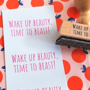 Puede incluir: Una tarjeta blanca con el texto "Wake up beauty, time to beast!" impreso en rosa. La tarjeta est&aacute; sobre un fondo rosa con un patr&oacute;n de c&iacute;rculos naranjas. Un sello de madera con el mismo texto est&aacute; encima de la tarjeta.