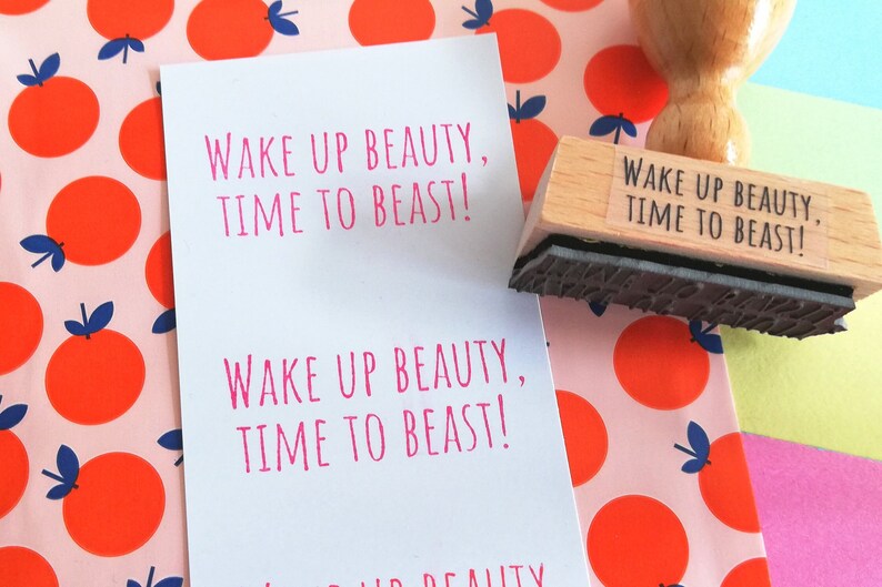 Puede incluir: Una tarjeta blanca con el texto "Wake up beauty, time to beast!" impreso en rosa. La tarjeta est&aacute; sobre un fondo rosa con un patr&oacute;n de c&iacute;rculos naranjas. Un sello de madera con el mismo texto est&aacute; encima de la tarjeta.