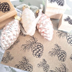 Könnte beinhalten: Eine Sammlung weihnachtlicher Artikel, darunter tannenzapfenförmige Ornamente in blassrosa mit goldenen Kappen, Holzstempel mit Tannenzapfenmotiven und eine Kraftpapierkarte mit einem sich wiederholenden Tannenzapfenmuster. Die Ornamente sind ca. 10 cm groß.