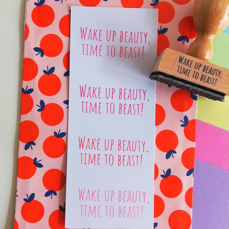 Puede incluir: Una tarjeta blanca con el texto "Wake up beauty, time to beast!" repetido cuatro veces. La tarjeta est&aacute; sobre un fondo rosa con un patr&oacute;n de naranjas rojas.