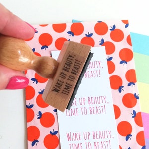 Puede incluir: Un sello de madera con el texto "Wake Up Beauty, Time To Beast!" impreso en &eacute;l. El sello est&aacute; siendo sostenido por una mano sobre un fondo estampado de color rosa y naranja.