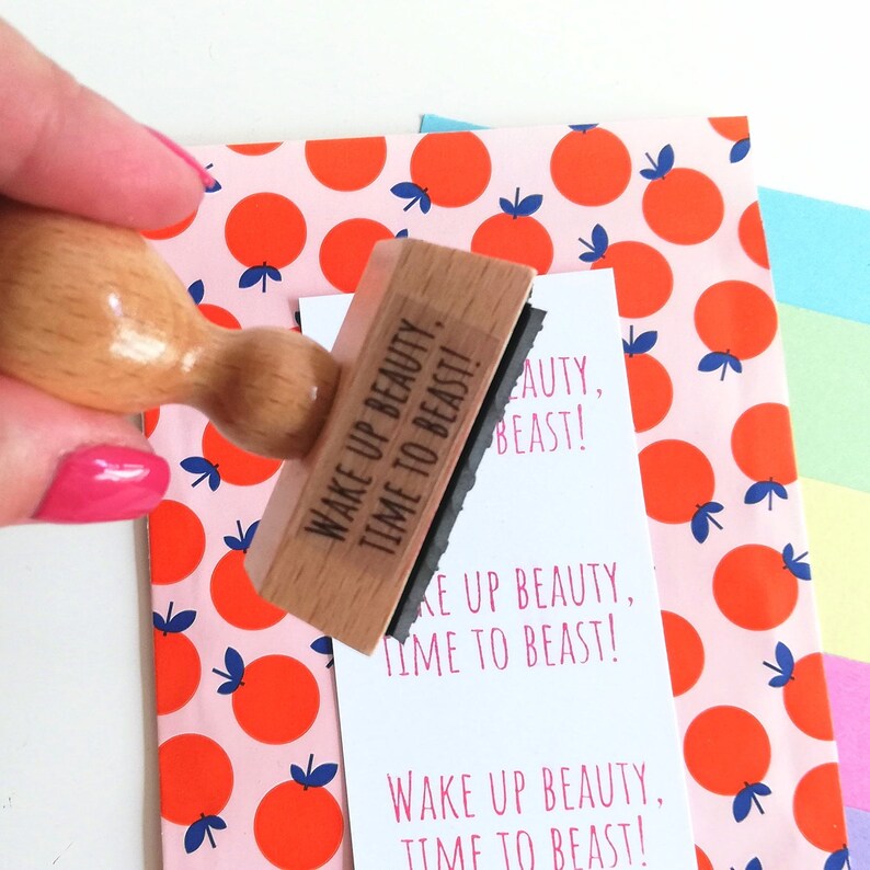 Puede incluir: Un sello de madera con el texto "Wake Up Beauty, Time To Beast!" impreso en &eacute;l. El sello est&aacute; siendo sostenido por una mano sobre un fondo estampado de color rosa y naranja.