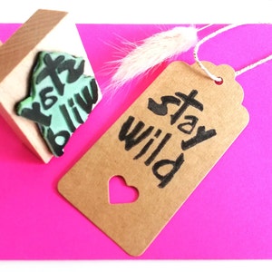 May include: A brown paper tag with a heart-shaped cutout and the text "Stay Wild" in black ink. The tag is attached to a white string and is lying on a pink surface. A wooden stamp with a green and black design is also visible in the image.