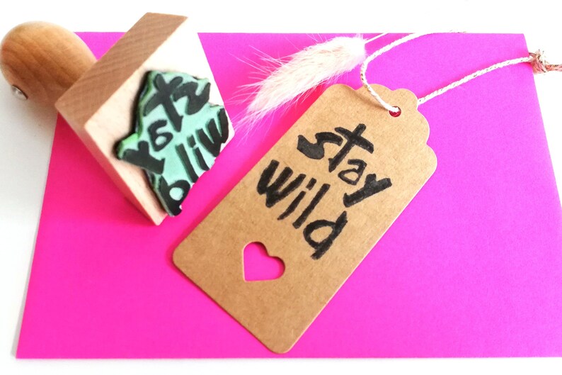 May include: A brown paper tag with a heart-shaped cutout and the text "Stay Wild" in black ink. The tag is attached to a white string and is lying on a pink surface. A wooden stamp with a green and black design is also visible in the image.