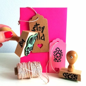 May include: Two wooden stamps with handles, one with a green and white design and the text "stay wild", the other with a black and white leopard print design. A pink card with a heart cut out and a spool of pink twine with gold accents.