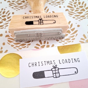 May include: A wooden stamp with the text "Christmas Loading" and a graphic of a progress bar with a gift box. The stamp is on a white background with a gold pattern.