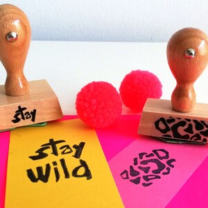 May include: Two wooden stamps with black ink. One stamp says "stay" and the other has a leopard print design. The stamps are on a pink and yellow surface with pink pom-poms.