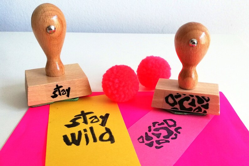 May include: Two wooden stamps with black ink. One stamp says "stay" and the other has a leopard print design. The stamps are on a pink and yellow surface with pink pom-poms.