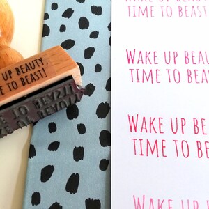 Puede incluir: Un sello de madera con el texto "Wake Up Beauty, Time To Beast!" impreso en &eacute;l. El sello est&aacute; sobre un fondo estampado azul y negro.