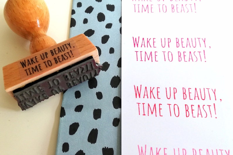 Puede incluir: Un sello de madera con el texto "Wake Up Beauty, Time To Beast!" impreso en &eacute;l. El sello est&aacute; sobre un fondo estampado azul y negro.