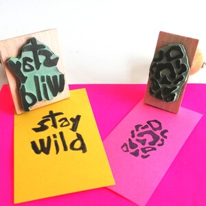 May include: Two wooden stamps with black ink. One stamp says "Stay Wild" and the other has a black and white abstract design. The stamps are on top of two pieces of paper, one yellow and one pink.