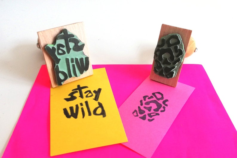 May include: Two wooden stamps with black ink. One stamp says "Stay Wild" and the other has a black and white abstract design. The stamps are on top of two pieces of paper, one yellow and one pink.