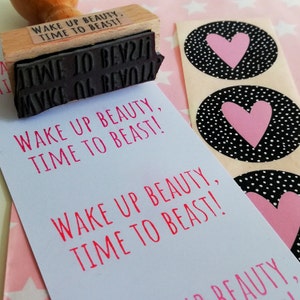 Puede incluir: Un sello de madera con el texto "Wake Up Beauty, Time To Beast!" en tinta negra. El sello est&aacute; sobre una tarjeta blanca con el mismo texto impreso en rosa.