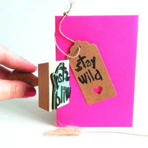 May include: A wooden stamp with the text "Stay Wild" in black ink on a light brown tag with a heart cutout. The tag is tied to a pink card with a string.