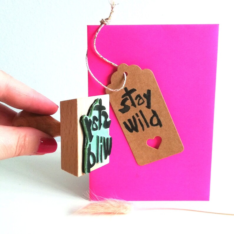 May include: A wooden stamp with the text "Stay Wild" in black ink on a light brown tag with a heart cutout. The tag is tied to a pink card with a string.