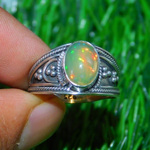 Può includere: Un anello d'argento con una pietra preziosa di opale di forma ovale. L'anello presenta un design dettagliato con piccole perle e una fascia testurizzata.