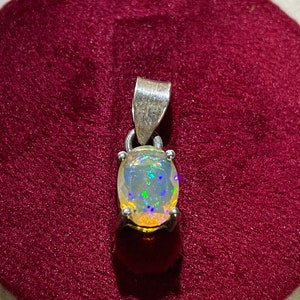 Puede incluir: Un colgante de plata con una piedra preciosa de ópalo blanco. El ópalo presenta un arcoíris de colores, incluyendo azul, verde y amarillo.