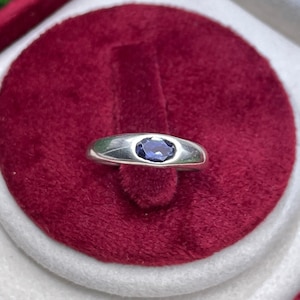 May include: A silver-toned metal ring featuring a single, oval-shaped, deep blue gemstone set flush into the center of the band. The band has a smooth, polished finish and a rounded profile, showcasing a clean and simple design with no additional ornamentation.