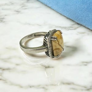 Peut inclure: Une bague en argent avec une grande pierre rectangulaire de couleur brun clair sertie dans une monture à griffes en argent. La pierre précieuse présente un motif marbré unique. La bague est posée sur une surface en marbre blanc.