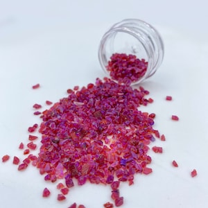 May include: A small clear jar with a lid is spilling out pink and red ruby gemstone chips onto a white surface.