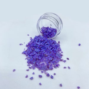May include: A small clear jar with purple iridescent glitter spilling out onto a white surface.