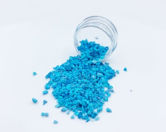 May include: A small glass jar with a blue, iridescent, crushed glass-like material spilling out onto a white surface.