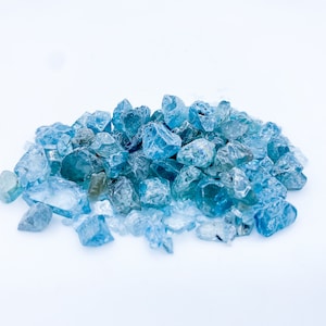 May include: A pile of light blue aquamarine gemstones, some with a slight green hue, on a white background.