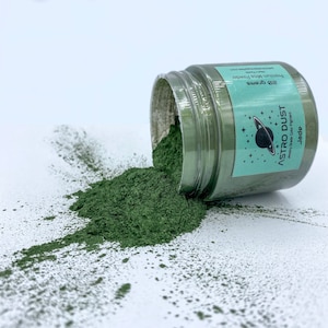 Puede incluir: Un tarro de polvo de mica verde Astro Dust Jade, que se derrama sobre una superficie blanca. El tarro está etiquetado con "Astro Dust" con un gráfico de planeta y estrellas. La etiqueta también dice "Pigmento de color de calidad para joyería" y "Jade". El tarro contiene 20 gramos de polvo.