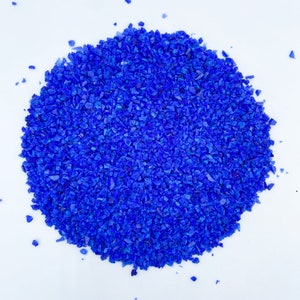 May include: A pile of small, irregularly shaped, vibrant blue glass pieces. The pieces are scattered on a white background, creating a textured, eye-catching display. The glass appears to be crushed or broken, with sharp edges and varying sizes.