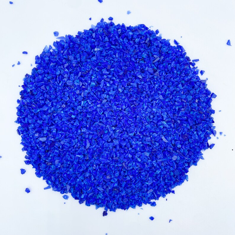 May include: A pile of small, irregularly shaped, vibrant blue glass pieces. The pieces are scattered on a white background, creating a textured, eye-catching display. The glass appears to be crushed or broken, with sharp edges and varying sizes.