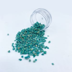 Peut inclure: Un pot en plastique transparent avec un couvercle déversant de l'opale concassée irisée turquoise. L'opale est dispersée sur une surface blanche.