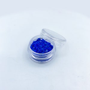 May include: A small, clear plastic jar filled with vibrant blue, crystalline fragments. The jar's lid is slightly ajar, revealing the contents. The background is a clean, white surface, highlighting the intense color of the crystals.