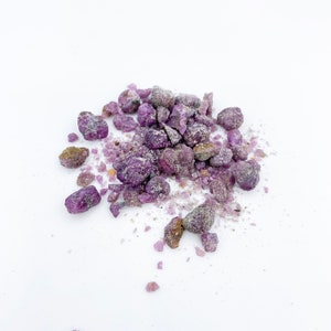 Può includere: Un mucchio di piccoli cristalli viola e grigi su uno sfondo bianco. I cristalli hanno una forma irregolare e una consistenza ruvida.