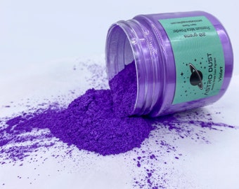 Puede incluir: Un tarro de polvo de mica púrpura etiquetado como "Astro Dust" con el texto "Premium Mica Powder" y "25 gramos". El tarro está abierto y el polvo se derrama sobre una superficie blanca.