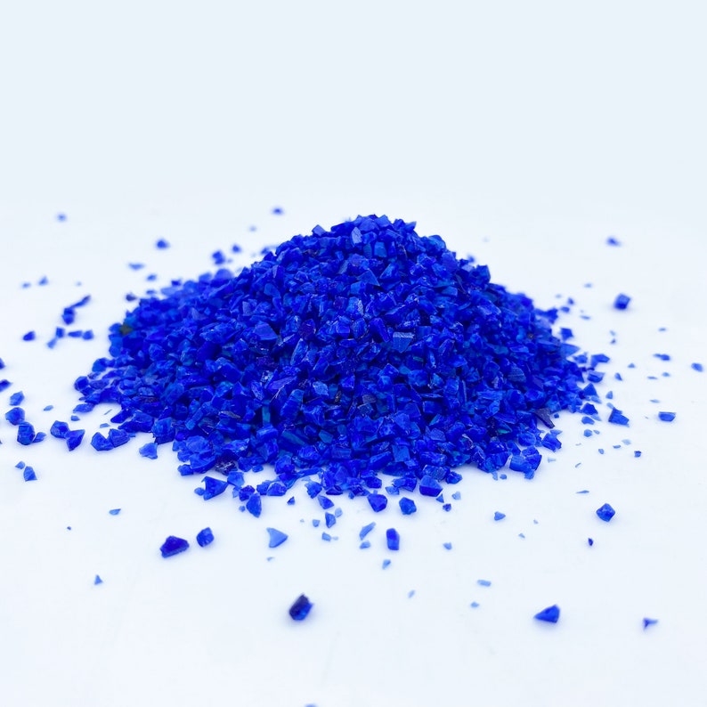 May include: A pile of vibrant, crushed blue glass crystals against a white background. The crystals are irregularly shaped and reflect light, creating a sparkling effect. The image is a close-up, highlighting the texture and color of the material.