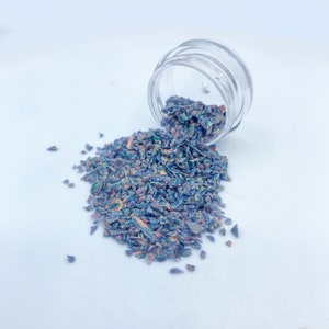 May include: A small jar of iridescent blue and purple glitter, with some of the glitter spilled out onto a white surface.